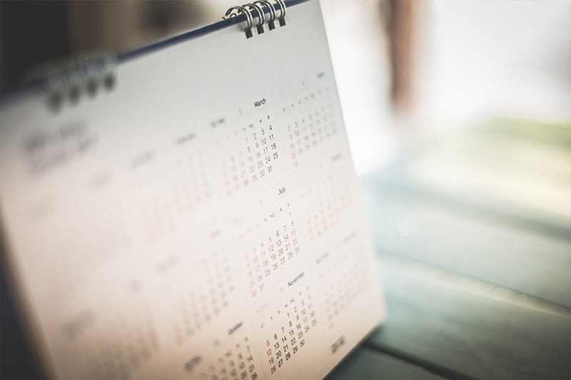 A close-up of a desk calendar displaying multiple months, with dates slightly blurred. The calendar rests on a wooden surface in soft, natural light.