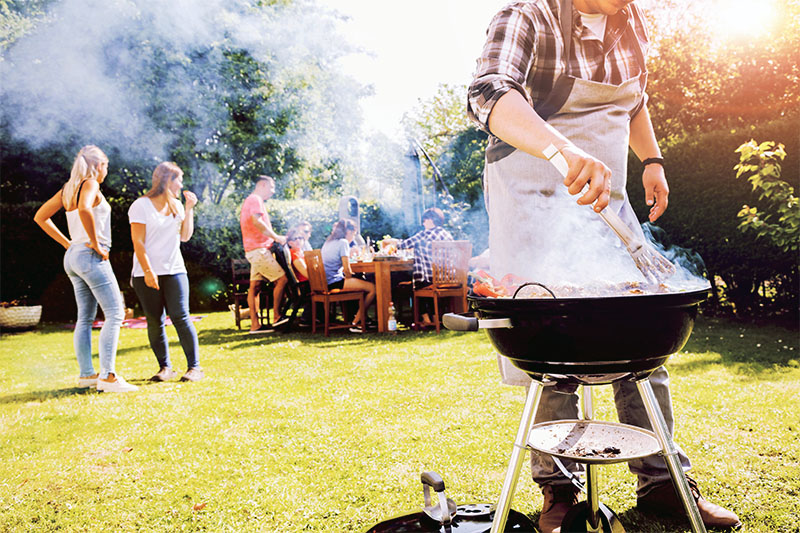 Home 3 A person grilling food on a barbecue in a sunny backyard while a group of people socialize and sit at a table in the background. Smoke rises from the grill, and trees and grass surround the scene.